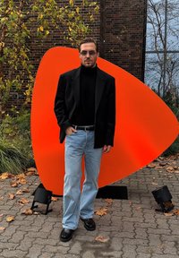 Blazer nero sopra un dolcevita nero, jeans azzurri, scarpe nere. Lo sfondo presenta una grande scultura arancione e foglie cadute sparse.