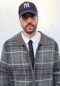 Veste en laine à carreaux verts, bleus et blancs avec fermeture éclair ; portée sur une chemise blanche et une cravate noire, assortie à une casquette de baseball foncée.