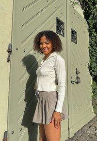 T-shirt court blanc à côtes avec des manches longues et un détail à cordon, associé à une jupe plissée beige, devant un fond de porte texturé vert.