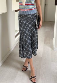 Jupe portefeuille à carreaux gris avec une coupe évasée en ligne A, associée à des talons noirs à brides. Accessoirisée d'une pochette noire ornée de strass.