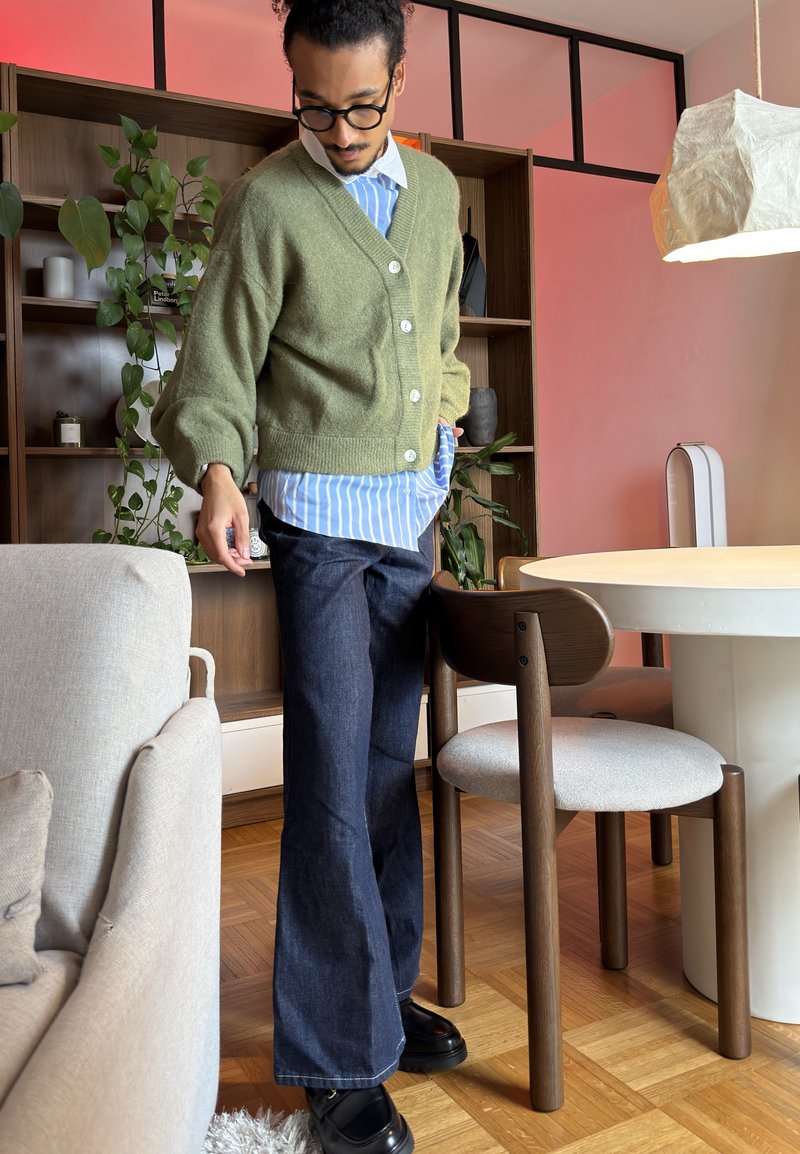 Silmälasipäinen ja kiharatukkainen mies seisoo modernissa olohuoneessa yllä vihreä neuletakki, sininen raidallinen paita, tummat leveälahkeiset farkut ja mustat kengät.