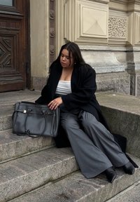 Siva kožna torba s zlatnim detaljima stoji pored sjedeće osobe u crnom kaputu, bijeloj majici i sivoj širokoj hlači na kamenim stepenicama.