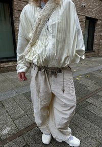 Balta klāta jaka, vaļīgas krēmkrāsas bikses ar sudraba šarmu jostu un gaišas slip-on kurpes, stāvot uz teksturēta bruģa.