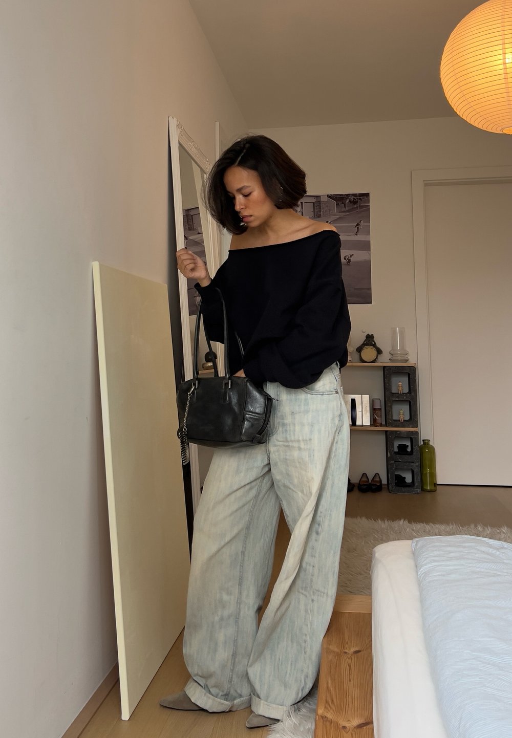 Femme en jean large clair et haut noir décolleté portant un sac à main noir, debout près d'un miroir mural dans une chambre avec un éclairage doux.