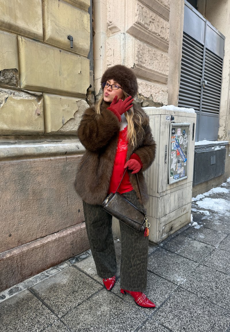 Femme en manteau de fourrure marron et chapeau, avec des gants rouges, un chemisier et des chaussures, posant sur un trottoir urbain enneigé près d'un mur de bâtiment usé.