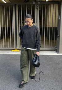 Oseba v temno sivi pleteni majici, olivno zelenih širokih hlačah in črnih čevljih drži črno torbo. Nosijo prozorna očala in minimalno nakit.