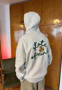 Personne portant un sweat à capuche gris clair avec le texte « state of mind » au dos, debout à l'intérieur près de placards en bois et d'une chaise verte.