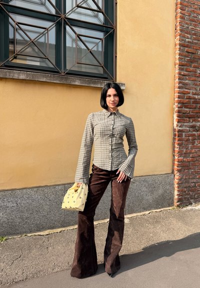 Mujer con cabello corto y negro, vestida con una camisa de cuadros de manga larga y pantalones oscuros acampanados, sosteniendo un bolso de mano amarillo pálido, de pie al aire libre.