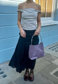 Off-the-shoulder ανοιχτό μπεζ τοπ, μαύρη πλισέ φούστα, μωβ τσάντα χειρός με διάφανα χερούλια, και γυαλιστερές μπορντό γόβες σε πεζοδρόμιο.