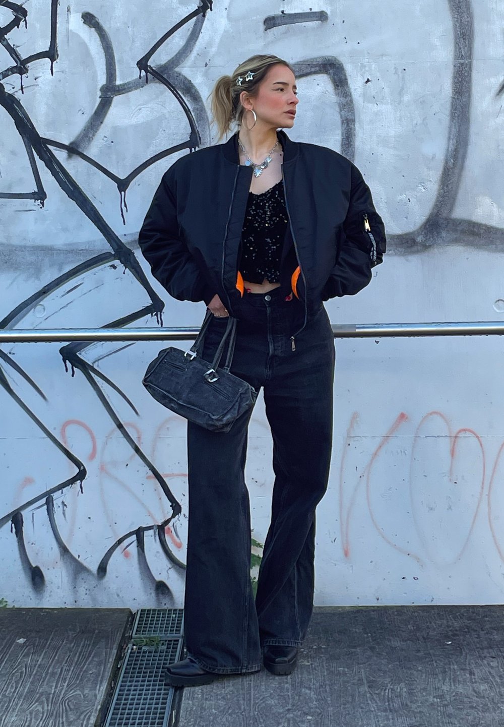 Μαύρο bomber μπουφάν με πορτοκαλί φόδρα, κροσέ τοπ, φαρδιά μαύρα τζιν παντελόνια και μια μικρή μαύρη τσάντα, μπροστά σε ένα γκράφιτι.
