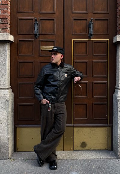 Chaqueta de cuero negra con cremallera, pantalones de pana marrón de pierna ancha y zapatos negros. De pie junto a puertas de madera con acentos dorados. Llavero adjunto.