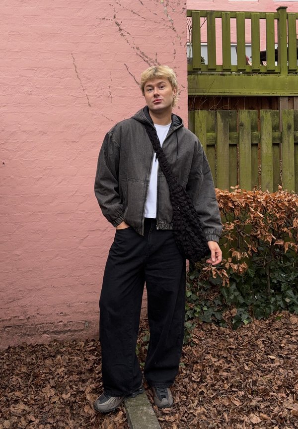 Νεαρός με σκούρο μπουφάν, φαρδιά μαύρη παντελόνα και τσάντα χιαστί, στέκεται σε έδαφος καλυμμένο με φύλλα κοντά σε ροζ τοίχο και πράσινο ξύλινο φράχτη.