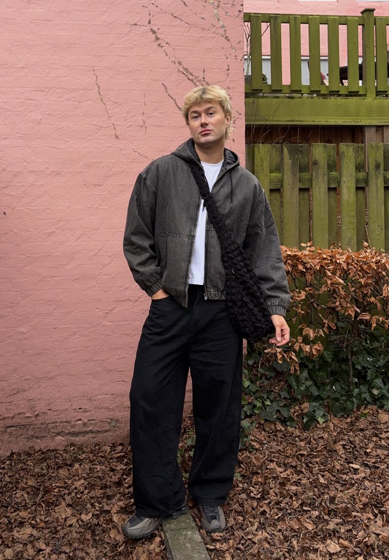 Jeune personne portant une veste sombre, un pantalon large noir et un sac bandoulière, debout sur un sol couvert de feuilles près d'un mur rose et d'une clôture en bois verte.