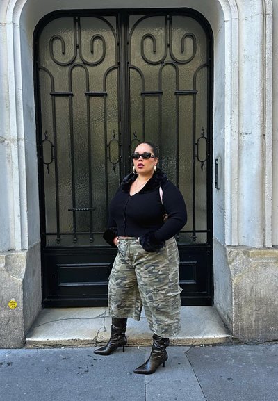 Cárdigan negro acanalado con puños de piel sintética, pantalones holgados de camuflaje y botas negras con tacón; de pie frente a una puerta de vidrio decorativa.