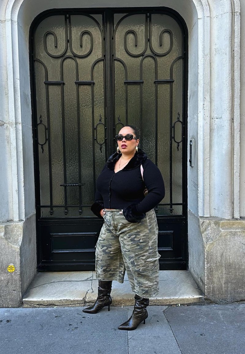 Cardigan noir à côtes avec poignets en fausse fourrure, pantalon baggy camouflage, et bottes noires à talons ; debout devant une porte en verre décorative.