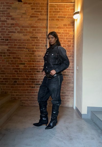 Chaqueta de mezclilla negra con bolsillos frontales, combinada con pantalones de mezclilla negra de corte holgado y botas de cuero negras hasta la rodilla. Fondo de ladrillo texturizado.