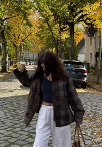 Bruine geruite jas met donkere accenten, witte drawstring broek en een marineblauw shirt. Houdt een koffiekop vast in een herfstachtige straatsetting.