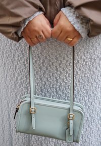 Χέρια με δαχτυλίδια που κρατούν τυρκουάζ τσάντα χειρός με χρυσές αγκράφες πάνω σε υφή λευκού υφάσματος και μπεζ μανίκια.