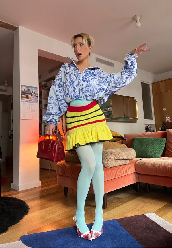 Blå blommig oversize zip-up topp, gul kjol med röda och svarta ränder, mintgröna strumpbyxor, röd handväska och blommiga höga klackar. Inomhusmiljö.