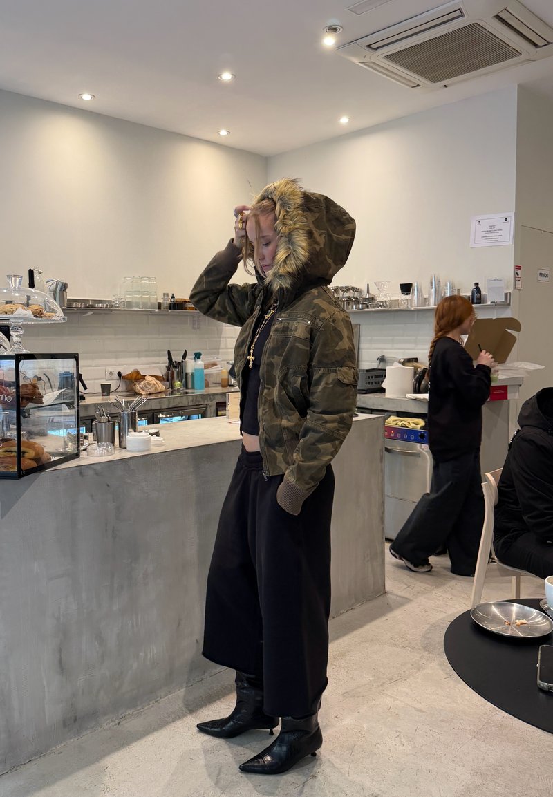 Personne portant une veste camouflage à capuche en fourrure, un haut noir court et un pantalon large, debout près d'un comptoir à café dans un café moderne.