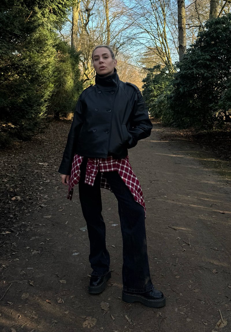 Une jeune personne se tient sur un sentier de terre dans une zone boisée, portant une veste noire, une chemise rouge à carreaux nouée autour de la taille, un pantalon noir et des chaussures à plateformes.