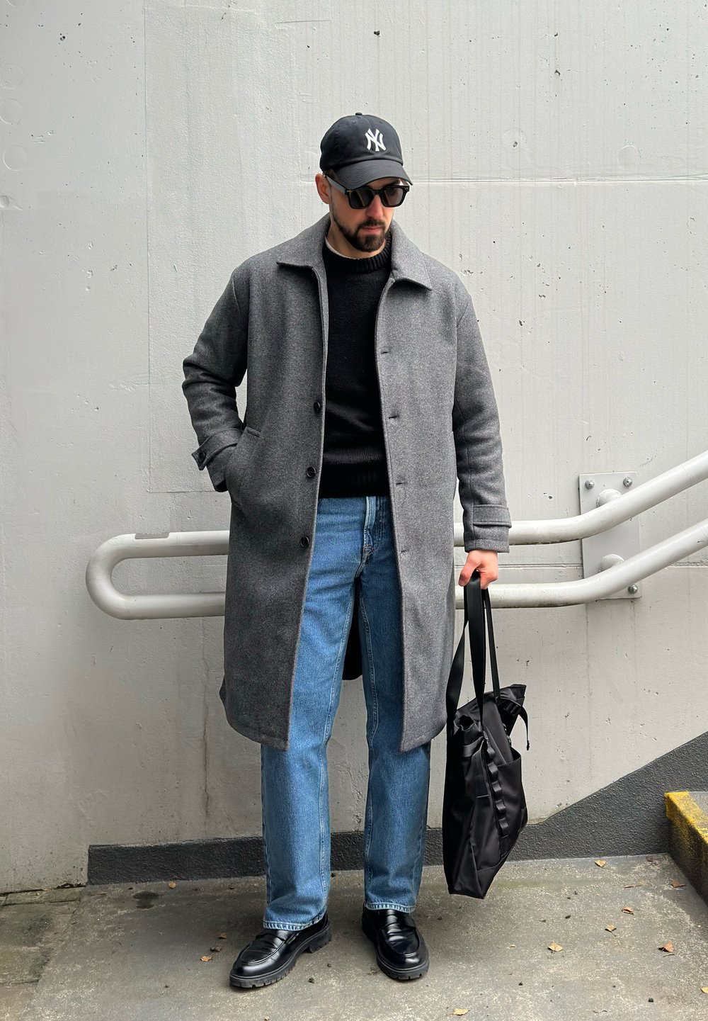 Manteau en laine gris, pull noir, jean bleu, chaussures noires, tenant un sac fourre-tout noir. Accessoires comprenant une casquette noire et des lunettes de soleil.