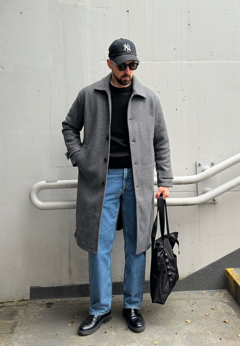 Manteau en laine gris, pull noir, jean bleu, chaussures noires, tenant un sac cabas noir. Accessoires comprennent une casquette noire et des lunettes de soleil.