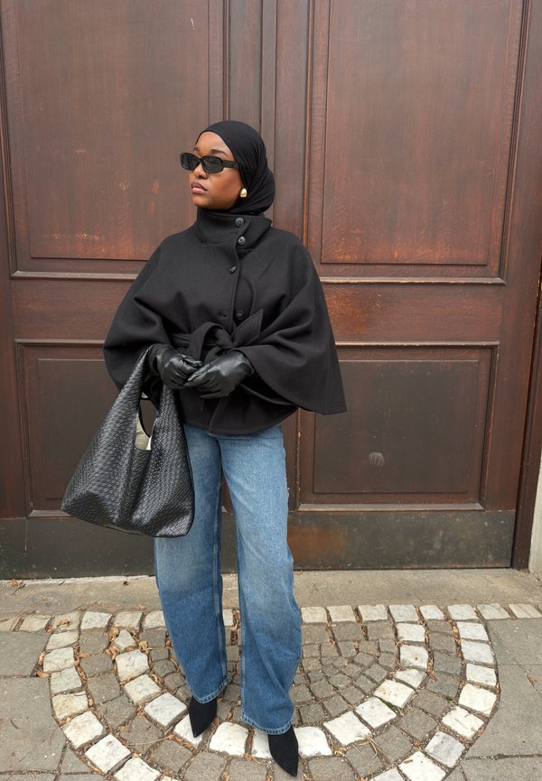 Kvinde iført sort hijab, solbriller, kappefrakke, læderhandsker, blå jeans og sorte støvler, holder en stor tekstureret sort håndtaske, står ved en trædør