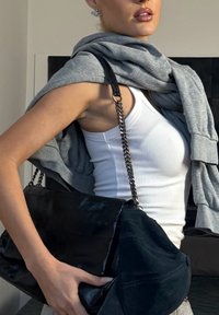 Gros plan sur une personne portant un débardeur blanc côtelé et un sweat-shirt gris posé sur les épaules, tenant un grand sac à main noir avec une bandoulière chaîne.