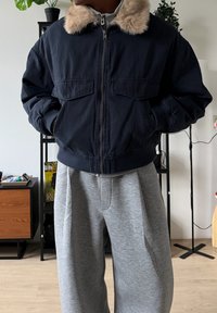 Veste aviateur marine avec col en fausse fourrure, fermeture éclair à l'avant et deux poches avant. Portée avec un pantalon de survêtement ample de couleur gris clair.