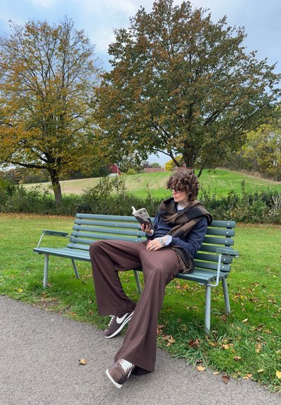 Una persona está sentada en un banco de parque verde, vistiendo una parte superior oscura, pantalones anchos marrones y zapatillas, leyendo un libro en un entorno al aire libre.