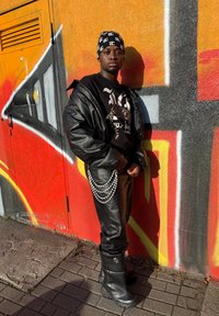 Sort læderjakke, sort t-shirt med grafik og sorte læderbukser med perlekæde. Sorte gummistøvler. Farverig graffiti som baggrund.