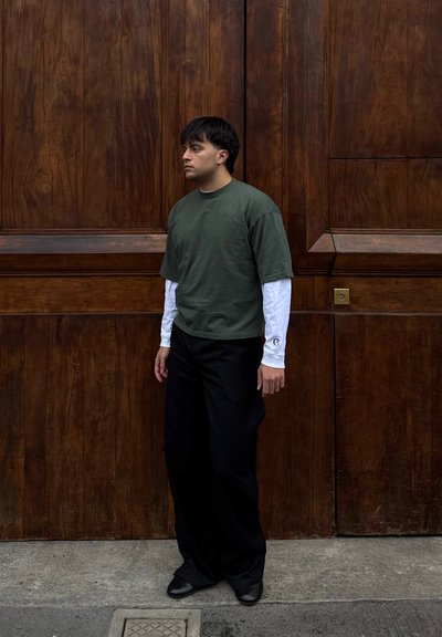 Hombre con camisa de manga corta verde oscuro sobre camiseta blanca de manga larga, pantalones negros, de pie en la acera frente a una gran puerta de madera.