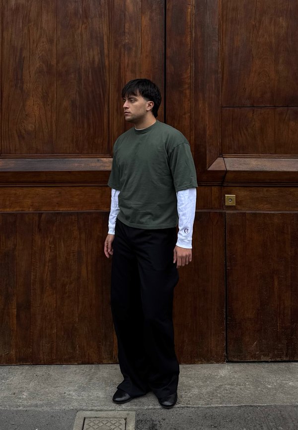 Muškarac u tamnozelenoj kratkoj majici preko bijelih dugih rukava, crne hlače, stoji na pločniku uz veliki drveni vrata u pozadini.