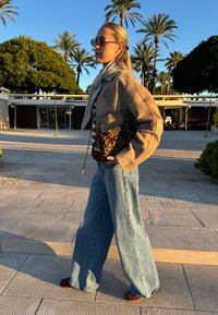 Veste en daim beige, haut rayé, jean large en denim, et sac à main à imprimé léopard. Lunettes de soleil et foulard complètent la tenue. Palmiers en arrière-plan.