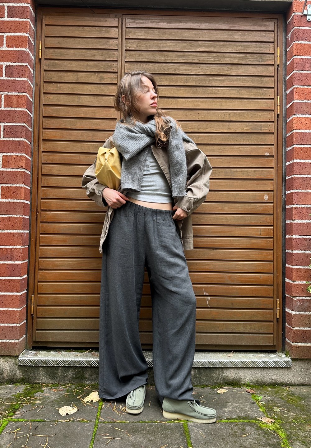 Grå bredbenede bukser, lys grå cropped top, oversized beige jakke og et gråt tørklæde, parret med grønne sko og en gul taske. Træbaggrund.
