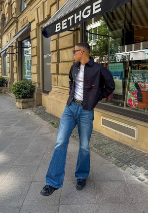Chaqueta negra de botones, camiseta blanca debajo, jeans de mezclilla azul de pierna ancha y zapatos negros. De pie en una acera con carteles de tiendas de fondo.