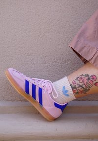 Scarpa sportiva rosa con strisce blu e suola in gomma, caratterizzata da una superficie liscia; indossata con una calza bianca e un tatuaggio floreale visibile.