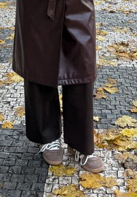 Brown leather apron over wide black trousers and grey sneakers with white laces, set against cobblestone pavement with fallen leaves.