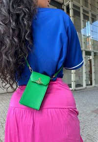Green leather phone pouch with gold hardware, suspended from a strap, worn against a blue top and pink skirt. Textured and rectangular design.