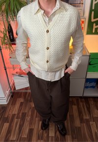 Kremfarget strikket vest med teksturert mønster og knappestengning, brukt over en stripete skjorte. Kombinert med løstsittende brune bukser og sko.