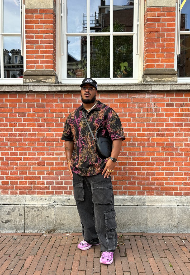 Μαύρο πουκάμισο κοντομάνικο με σχέδια ροζ και χρυσά κυκλώματα, συνδυασμένο με μαύρο παντελόνι cargo και ροζ αθλητικά παπούτσια. Το αξεσουάρ περιλαμβάνει μια μαύρη τσάντα χιαστί.