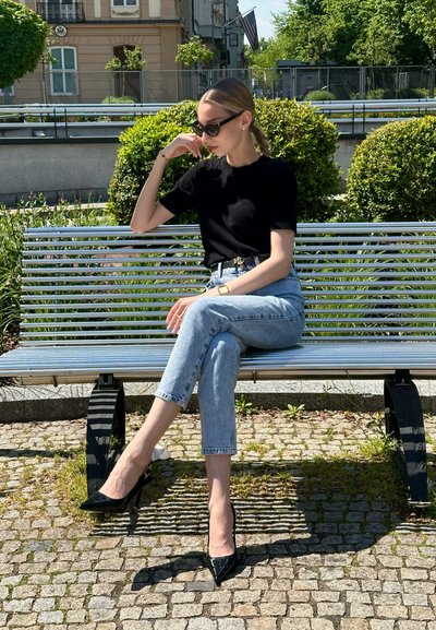 Camisa negra de manga corta, jeans cortos de mezclilla azul claro y tacones de charol negros. Persona sentada en un banco plateado en un área verde al aire libre.