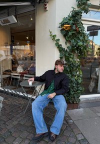 Persona seduta a un piccolo tavolo rotondo in metallo, indossa una giacca nera, una maglietta verde e jeans blu larghi, tiene in mano una tazza. L'edera cresce sul muro.