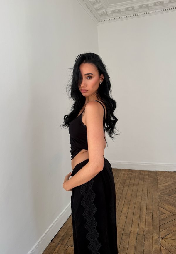 Jeune femme aux longs cheveux noirs portant un crop top noir et un pantalon large, debout de côté dans une pièce avec un sol en bois et des murs blancs.