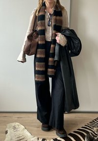 Braune Lederhandtasche, gemusterter schwarzer, brauner und grüner Schal, hellbeige Oberteil, schwarze weite Hose, schwarzer Mantel, braune Stiefel mit strukturierten Sohlen.