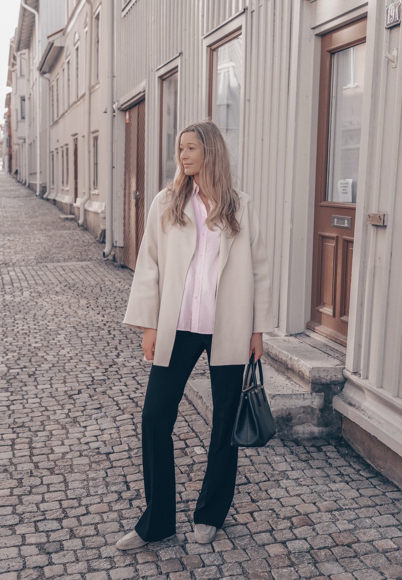 Femme en manteau clair, chemise rose, pantalon noir, chaussures grises, tenant un sac à main noir, debout sur une rue pavée à côté de bâtiments de couleur claire.