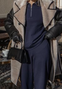 Casaco longo de pelo bege e preto, top azul-marinho, calças escuras e luvas. Bolsa preta com detalhe de fecho dourado. Ambiente de café ao ar livre.