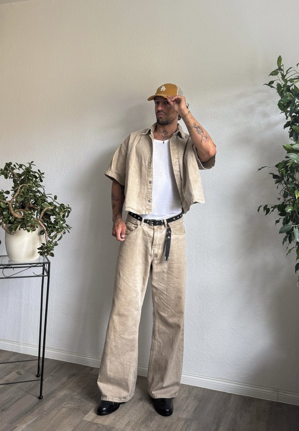 Man draagt beige broek, open kort mouwhemd, wit hemd, zwarte riem en gele pet, staat tussen twee groene kamerplanten binnen.