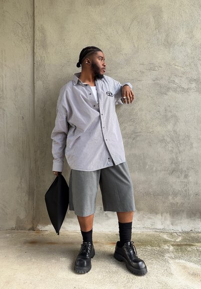 Camisa de botones de gran tamaño con rayas suaves y un logo, combinada con pantalones cortos grises hasta la rodilla. Botas negras con cordones y una bolsa negra.